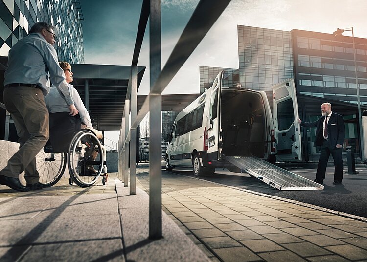 Una persona in sedia a rotelle viene accompagnata verso un minibus con la rampa di accesso abbassata.