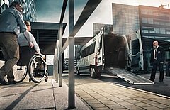 Ein Rollstuhlfahrer wird zu einem KMP-Fahrzeug mit ausgeklappter Rampe begleitet.
